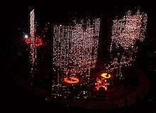 A darkened stage seen from above. Curtains of red and white lights hang down behind and to the sides of the four band members who are lit up with spotlights. Three of the members are on the main stage while the fourth is on the b-stage, an elliptical catwalk that surrounds the main stage.