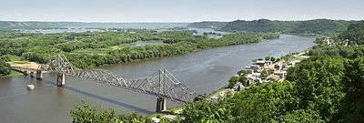 Summertime overlooking a wide and winding river.  A small town is to the right.  An old steel cantilever-truss bridge dominates the foreground.