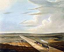 A very early steam locomotive pulling four open carriages under a cloudy blue sky along a track slightly built up from the surrounding flat countryside. The train has just passed a small farmhouse and is approaching a gentleman who is standing by the side of the track.