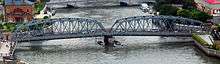 A bridge of two gray metal spans, curving segmentally above, crossing a river, seen from an upward angle