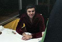 A caucasian man wearing a dark burgandy shirt sits at a table.