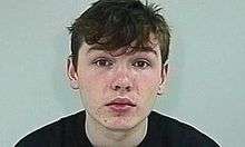 A white male teenager with dark hair and a black t-shirt, shown from the shoulders up. He has a neutral expression and is against a grey backdrop.