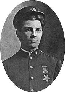 Head and shoulders of a young white man wearing a cap pushed high up on his forehead and a plain military jacket with a star–shaped medal hanging from a ribbon pinned to his left breast.