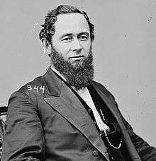 A man with curly black hair and a beard wearing a black jacket and vest and a white shirt