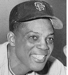 The face of a dark-skinned man who is smiling widely. The letters "S" and "F" overlap on his hat.