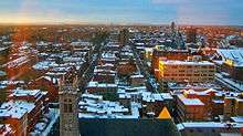 Several city blocks seen from above near a setting sun off-camera to the left, with snow on their roofs and some church spires sticking up