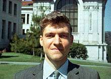 A man in a suit faces the camera while he stands in front of a building.