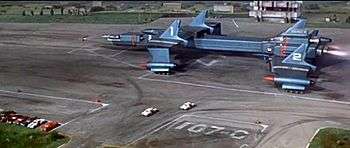 A massive, blue-grey spacecraft, roughly shaped like a plane but composed of multiple components, is on a runway surrounded by vehicles and buildings at the point of take-off.